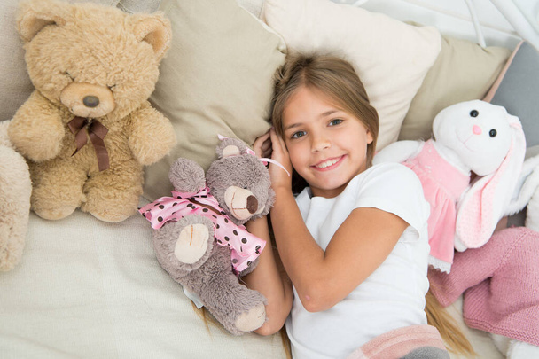 Comfortable bedroom. Relaxing before sleep. Girl enjoy evening time with toys. Kid lay bed with toys pillow blanket background. Girl child wear pajamas. Play soft toy before go sleep. Sleep with toy - Fotografie, Obrázek