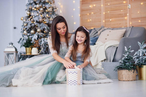 Beautiful mother and her cute daughter unpacking holidays gift while sitting on the floor near the Christmas tree in their cozy home. Happy winter holidays concept. - Фото, зображення