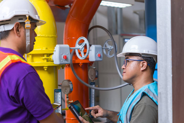 zwei asiatische Ingenieure, die im Heizungsraum arbeiten, die technischen Daten der Heizungsanlage überprüfen, thailändische Leute, diskutieren das Problem des Rohrdrucks. - Foto, Bild