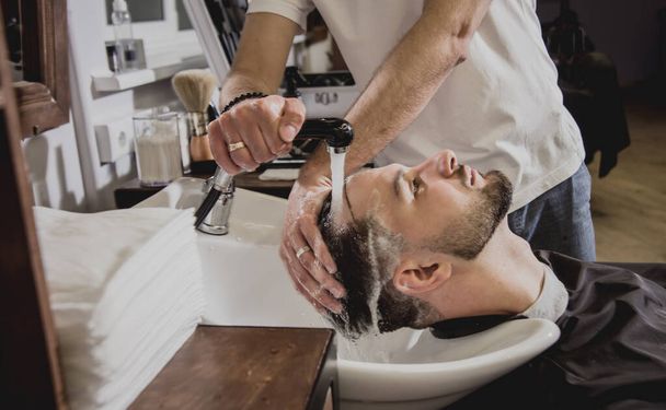 Young man with trendy haircut at barber shop. Barber washes customer head. Concept barbershop. - 写真・画像