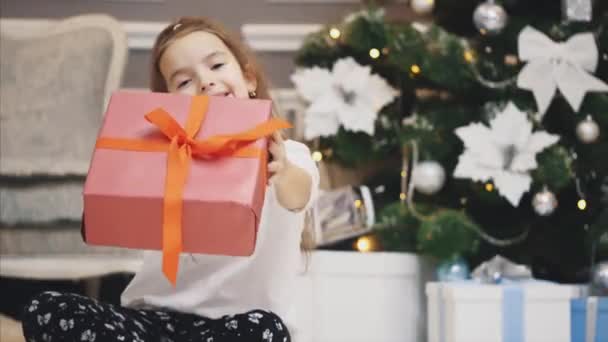 Closeup advertising video where little girl got a giftbox and presents it in front of the camera on Christmas background. - Video