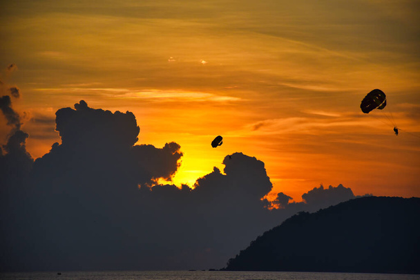 erstaunliche, wunderbare und dramatische goldfarbene Himmel hinter den Wolken während eines atemberaubenden Sonnenuntergangs, während Tandemparasailer den magischen Moment auf der Insel Langkawi in Malaysia erleben - Foto, Bild