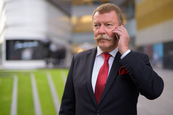 Όμορφος ανώτερος επιχειρηματίας με μουστάκι που χρησιμοποιεί το τηλέφωνο στην πόλη - Φωτογραφία, εικόνα