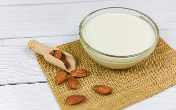 Almond milk in bowl and almonds on wooden scoop on sack  - Photo, image