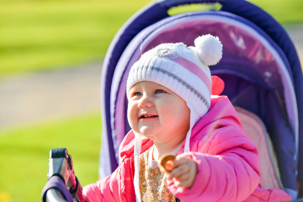Kırmızı ceketli, beyaz şapkalı küçük neşeli bir kız sonbahar parkında bebek arabasına biniyor.. - Fotoğraf, Görsel