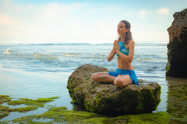 Fiatal nő ül a sziklán, meditál, jógázik és pranayama a namaste mudra a strandon, Bali - Fotó, kép