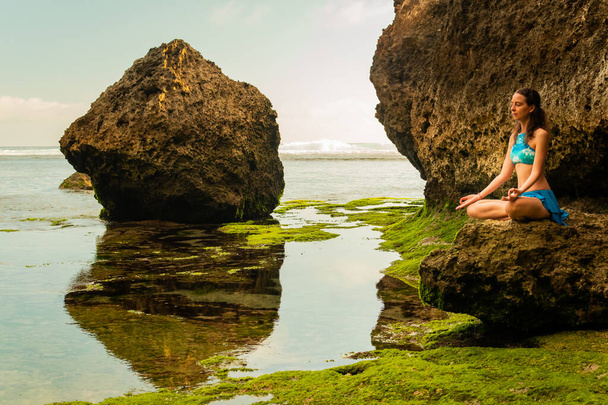 Jovem mulher sentada na rocha, meditando, praticando ioga e pranayama na praia, Bali. Mãos em mudra gyan
. - Foto, Imagem