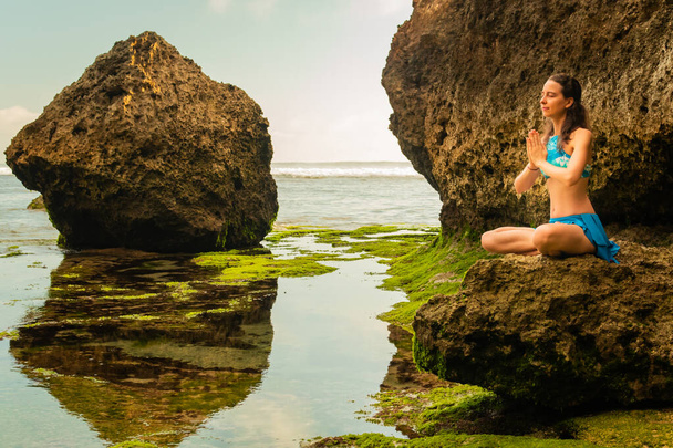 Mujer joven sentada en la roca, meditando, practicando yoga y pranayama en la playa, Bali. Manos en namaste mudra
 - Foto, imagen