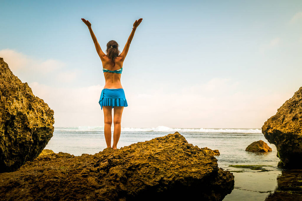 Excited young woman raising arms at the beach in front of the ocean. View from back. Sunset at the beach. Bali, Indonesia. - Photo, Image