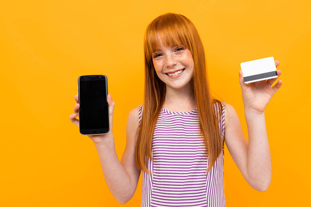Pretty teenage girl with red hair posing with mobile phone against orange background  - 写真・画像
