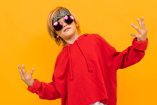 Netter modischer Junge mit rotem Kapuzenpulli und Sonnenbrille posiert gegen Orange  - Foto, Bild