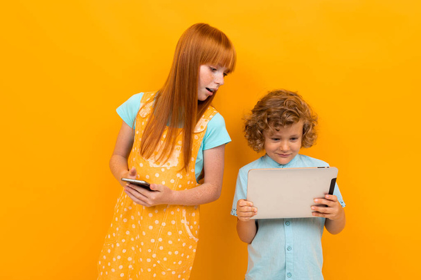 Mooi tiener meisje met rood haar en kleine jongen poseren tegen oranje achtergrond  - Foto, afbeelding