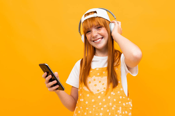 hübsches Teenager-Mädchen mit roten Haaren posiert mit Handy und Kopfhörern vor orangefarbenem Hintergrund  - Foto, Bild