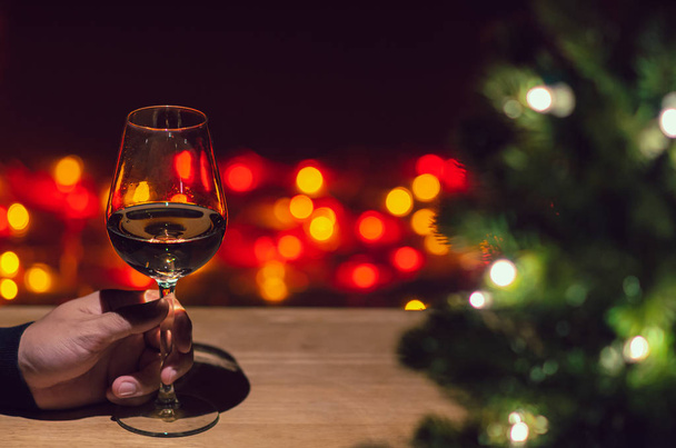 Hand toasting a glass of Rose wine on wooden table with Christmas tree and colorful bokeh light background. - Foto, Imagem