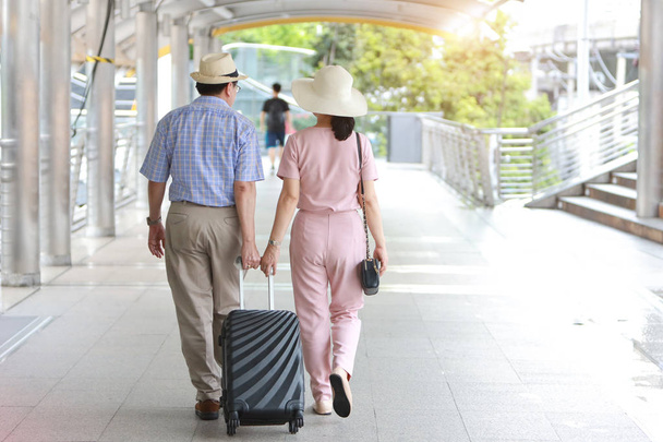 Вид сзади Азиатский путешественник пара муж и жена в розовой и синей рубашке с белой красивой шляпой и багажом ходьба в центре города с счастливым улыбающимся лицом во время пенсионного возраста - Фото, изображение