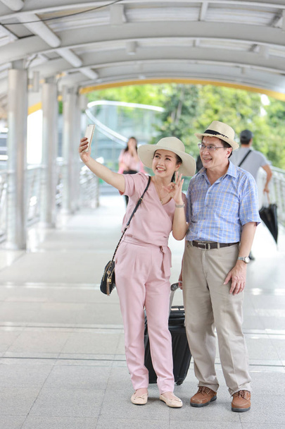 full length asian traveler couple husband and wife in pink and blue shirt with white nice hat and luggage walking in downtown with happy smiling face and taking photo from mobile while showing victory symbol  - Photo, Image