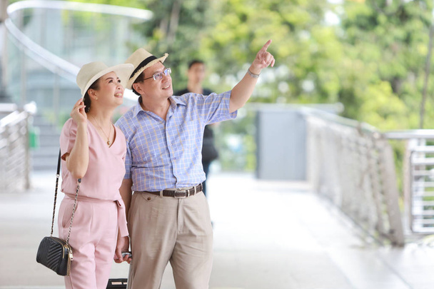 Azji podróżnik para mąż i żona w różowy i niebieski koszula z biały ładny kapelusz i bagaż spaceru w centrum miasta, on wskazując coś z prawej przestrzeni kopia - Zdjęcie, obraz