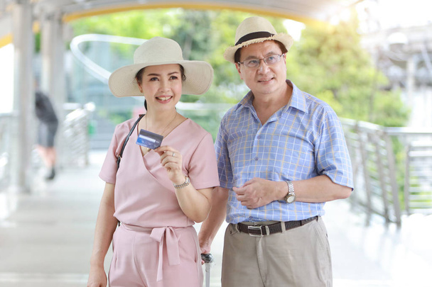 asiatisches Reisepaar Mann und Frau in rosa und blauem Hemd mit weißem Hut und Gepäck spaziert mit glücklich lächelndem Gesicht durch die Innenstadt und zeigt Kreditkarte für Einkäufe im Rentenalter - Foto, Bild