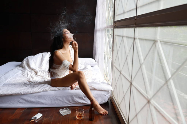 side view drunken asian woman in white lingerie, drinking and smoking while holding bottle of liquor alcohol and sitting on bed in bedroom - Photo, image