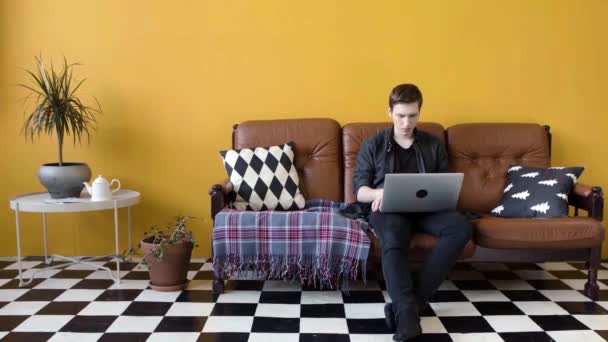 Egy fiatalember, aki otthon dolgozik a laptopon. Készletfelvétel. A fickó otthon dolgozik, mint szabadúszó a laptopon ül a kanapén. Modern munkastílus otthon szabadúszó módban - Felvétel, videó