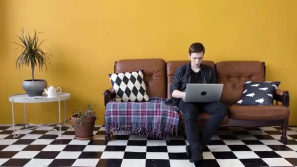Egy fiatalember, aki otthon dolgozik a laptopon. Készletfelvétel. A fickó otthon dolgozik, mint szabadúszó a laptopon ül a kanapén. Modern munkastílus otthon szabadúszó módban - Felvétel, videó