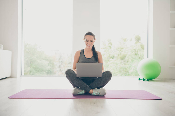 Full body photo of positive sportive girl crossing her legs sit on violet mat use laptop browse films movie about sport practicing in gym like house indoors - Fotografie, Obrázek
