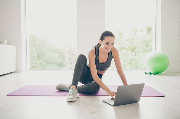 Full body photo of positive sporty girl using her computer browsing internet want find film movies video about sport fitness sit on purple mat in studio like house indoors - Фото, зображення