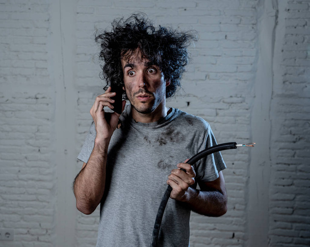 Portrait of young man holding electrical cable after domestic accident with dirty burnt funny face expression calling desperate with mobile phone asking for help. Electricity repairs and DIY concept. - Fotoğraf, Görsel