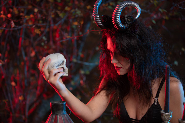 piękna wiedźma z rogami na głowie robi czarną magię na ludzkiej czaszce na święto halloween. Czarownica halloween kostium. - Zdjęcie, obraz