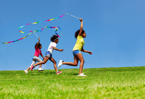 Jumping happy three girls wave with ribbon over blue sky running on grass - Фото, изображение