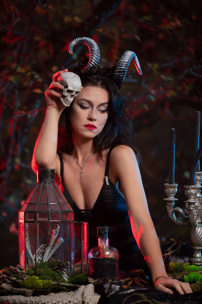 mooie heks met hoorns op haar hoofd die zwarte magie doet op een menselijke schedel voor Halloween viering. Witch Halloween kostuum. - Foto, afbeelding