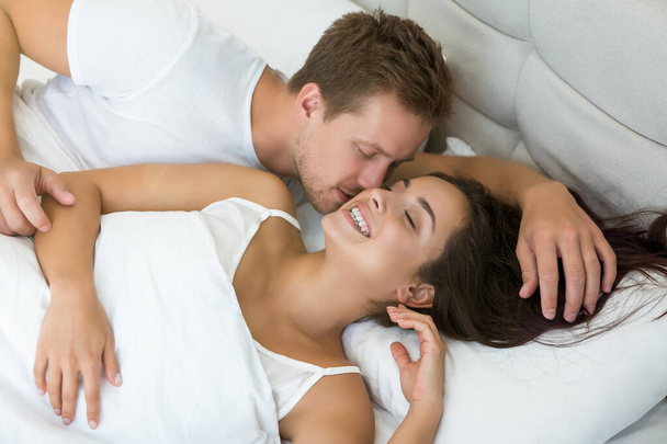 young couple handsome husband kissing his beautiful wife cuddling in bed in bright bedroom smiling to each other feeling happy family morning idyll - 写真・画像