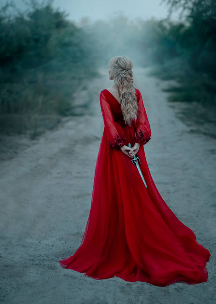 dangerous blonde queen in red fashion lush dress Hides a dagger behind. Backdrop dark fantasy forest in fog. Concept Revenge conspiracy betrayal. Halloween party. Turned away without face. Long train - Foto, imagen