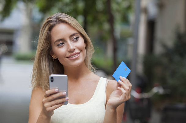 Smiling woman holding credit card and smart phone thinking looking at side in the street - Фото, изображение