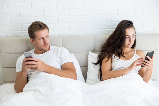 young couple handsome husband and his beautiful wife just woke up in bed checking their smartphones early in the morning man looks suspective social media addiction - Foto, afbeelding