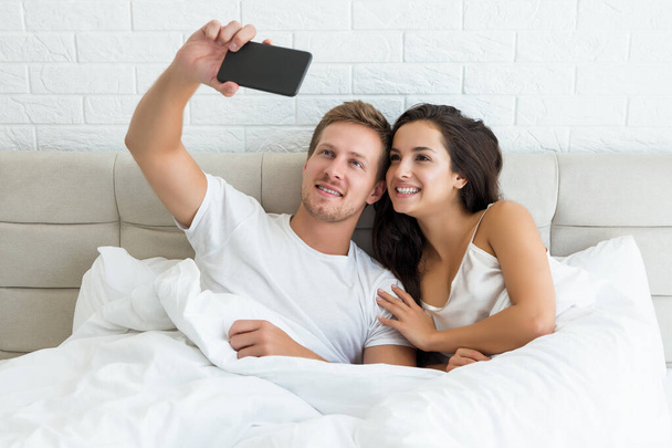 молода щаслива пара красивий чоловік і його красива дружина щойно прокинулася в ліжку, знімаючи селфі на смартфоні рано вранці залежність від соціальних мереж
 - Фото, зображення