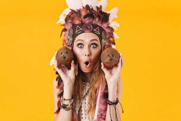 Portrait of beautiful surprised woman in headdress of feathers holding coconuts and looking at camera - Photo, Image
