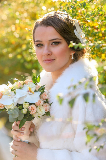 A noiva em um casaco de pele branco em um fundo de arbustos mantém um buquê de casamento em suas mãos
. - Foto, Imagem