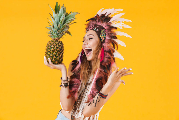 Portrait of beautiful delighted woman in headdress of feathers holding pineapple and laughing - Фото, изображение