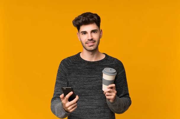 Young man over isolated background holding coffee to take away and a mobile - Photo, Image