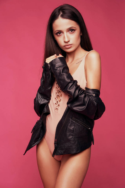 Model in pink bodysuit. Graceful young woman in black jacket standing isolated on bright background - 写真・画像