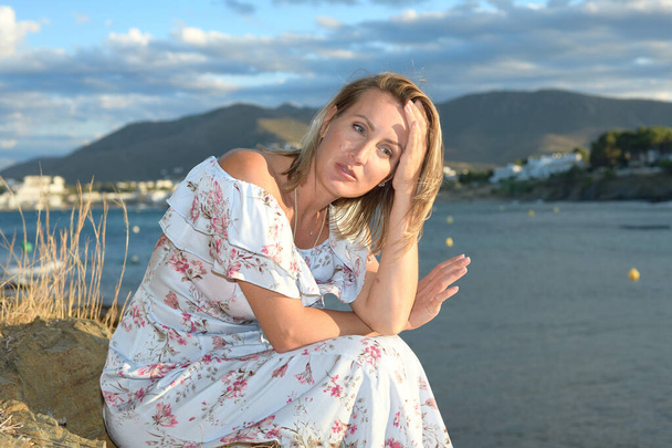 Nainen istuu pohjoisen Välimeren rannikkomaiseman edessä kesäiltapäivänä tuulen ja kopiotilan kanssa.
 - Valokuva, kuva