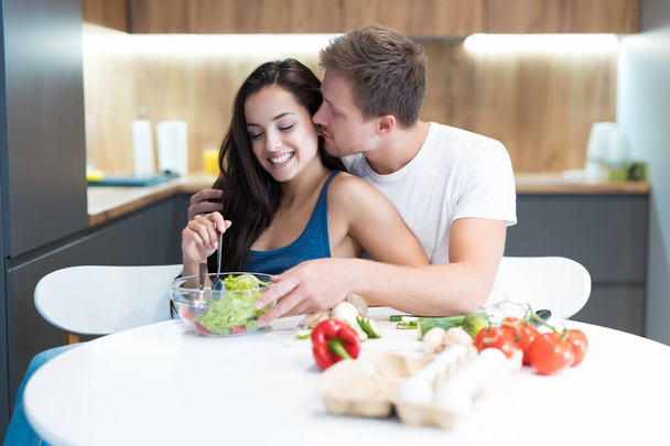 νεαρό ζευγάρι μαγειρεύουν πρωινό στην κουζίνα μαζί ο σύζυγος αγκαλιάζει και φιλάει την αγαπημένη του γυναίκα ενώ αναμιγνύει τις οικογενειακές παραδόσεις σαλάτας - Φωτογραφία, εικόνα