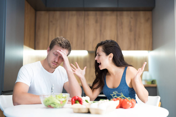 young couple having fight while cooking breakfast in the kitchen wife shouts loud at her husband while he looks annoyed family quarrel - Fotoğraf, Görsel