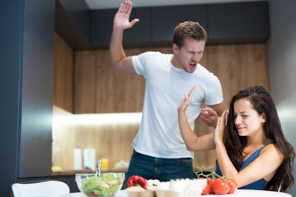 young couple having fight while cooking breakfast in the kitchen husband lifts his arm against his wife while she turns away domestic abuse - Fotografie, Obrázek