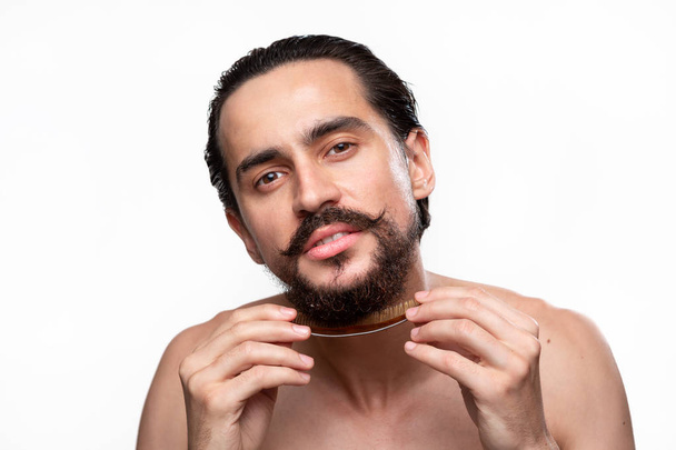 Elegante guapo barbudo con bigotes cepillándose la barba aislada sobre fondo blanco. Concepto de tratamiento matutino. Rutina matinal
 - Foto, imagen