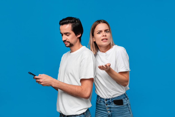 Μπρουνέ άντρας σε λευκό μπλουζάκι και μπλε τζιν κρατώντας το κινητό τηλέφωνο απομονωμένο πάνω από το μπλε φόντο. Κορίτσι προσπαθεί να δει τι είναι σε μια οθόνη ενός smartphone. - Φωτογραφία, εικόνα