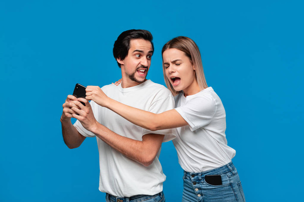 Hombre Brunet en camiseta blanca y jean azul sosteniendo teléfono móvil aislado sobre fondo azul. Chica trata de agarrarlo y comprobarlo
. - Foto, Imagen