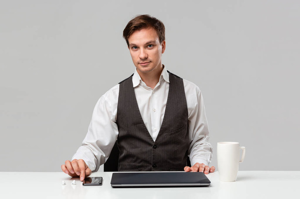 Pretty office worker in a white t-shirt and grey vest putting things in order at his working area. Laptop, smartphone and cup of coffee on a white table. - Photo, Image
