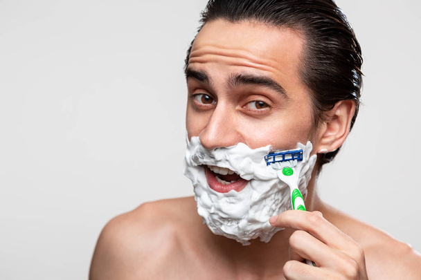 Красивий чоловік з вусами використовує палицю для гоління та піну для гоління, щоб обрізати свою бороду, що стоїть оголеною, ізольовано на білому тлі. Концепція ранкового лікування та гоління
. - Фото, зображення